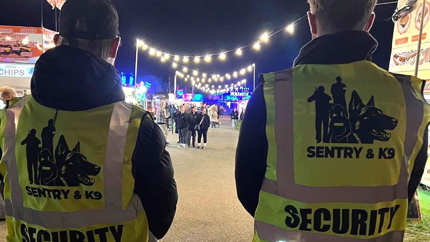 Two security guards stand with their backs to the camera, facing towards a walkway with rows of food kiosks. String lights zig zag across the walkway and small groups of people are seen in teh distance. Further away are bright blue and pink lights.