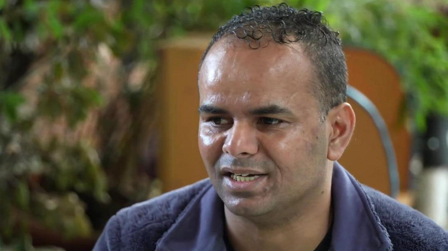 Mohamed has short black curly hair and stubble. He is wearing a blue jumper and is speaking to a BBC reporter interviewing him, who is out of shot.
