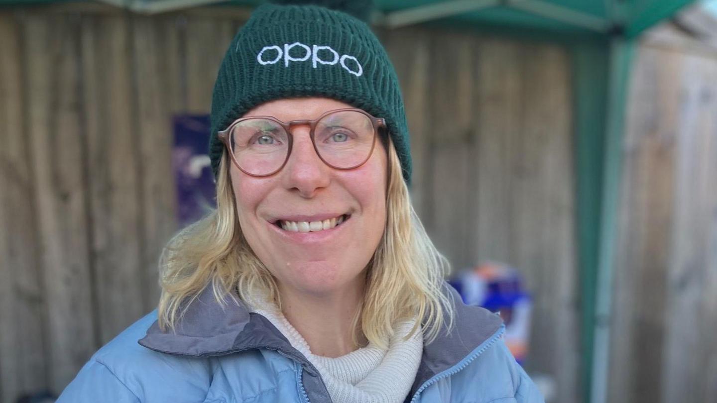 Janine pictured in her wheelchair and holding a charity donation bucket. She has shoulder length blonde hair and is wearing a blue coat and dark blue hat. 