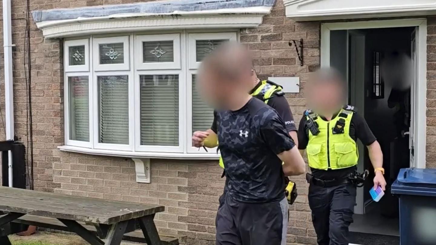 One of the suspects being led out of a home in Washington by police. His hands are handcuffed behind his back and he is being held by one of the officers. Another officer is following behind holding a notebook.