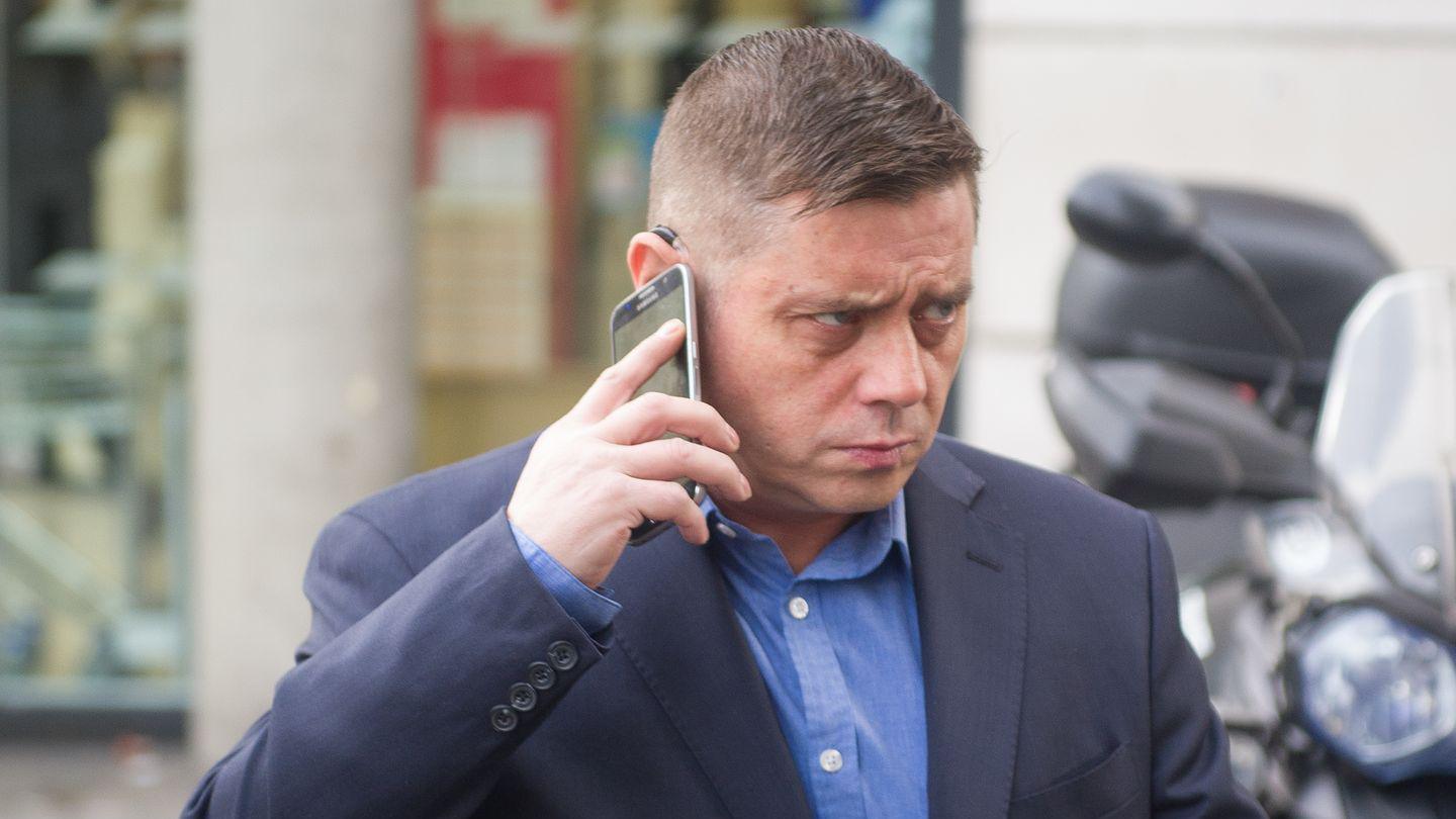 John Alford is photographed outside, and he has a mobile phone pressed to his ear and is walking forwards, not looking at the camera. He is frowning and has his lips pursed. He wears a blue shirt and navy blue suit jacket.