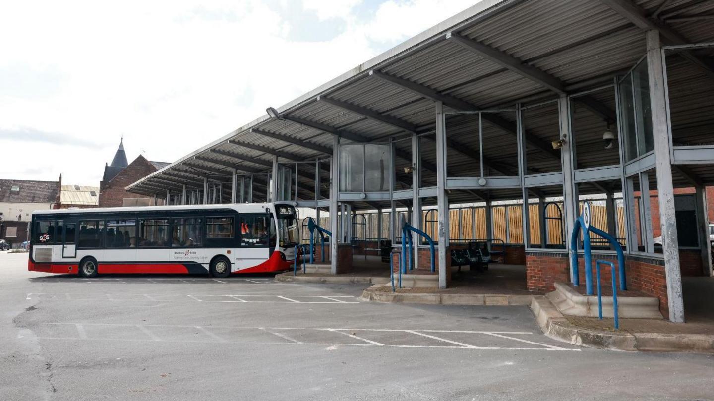 A white and red bus at Longton bus station which has several stops visible with a canopy above it. 
