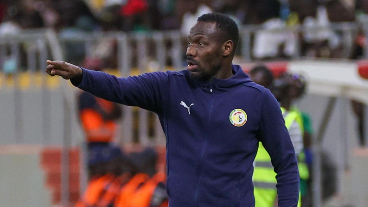 Pape Thiaw, wearing a purple Senegal tracksuit top, points with his right arm from the touchline at a football match