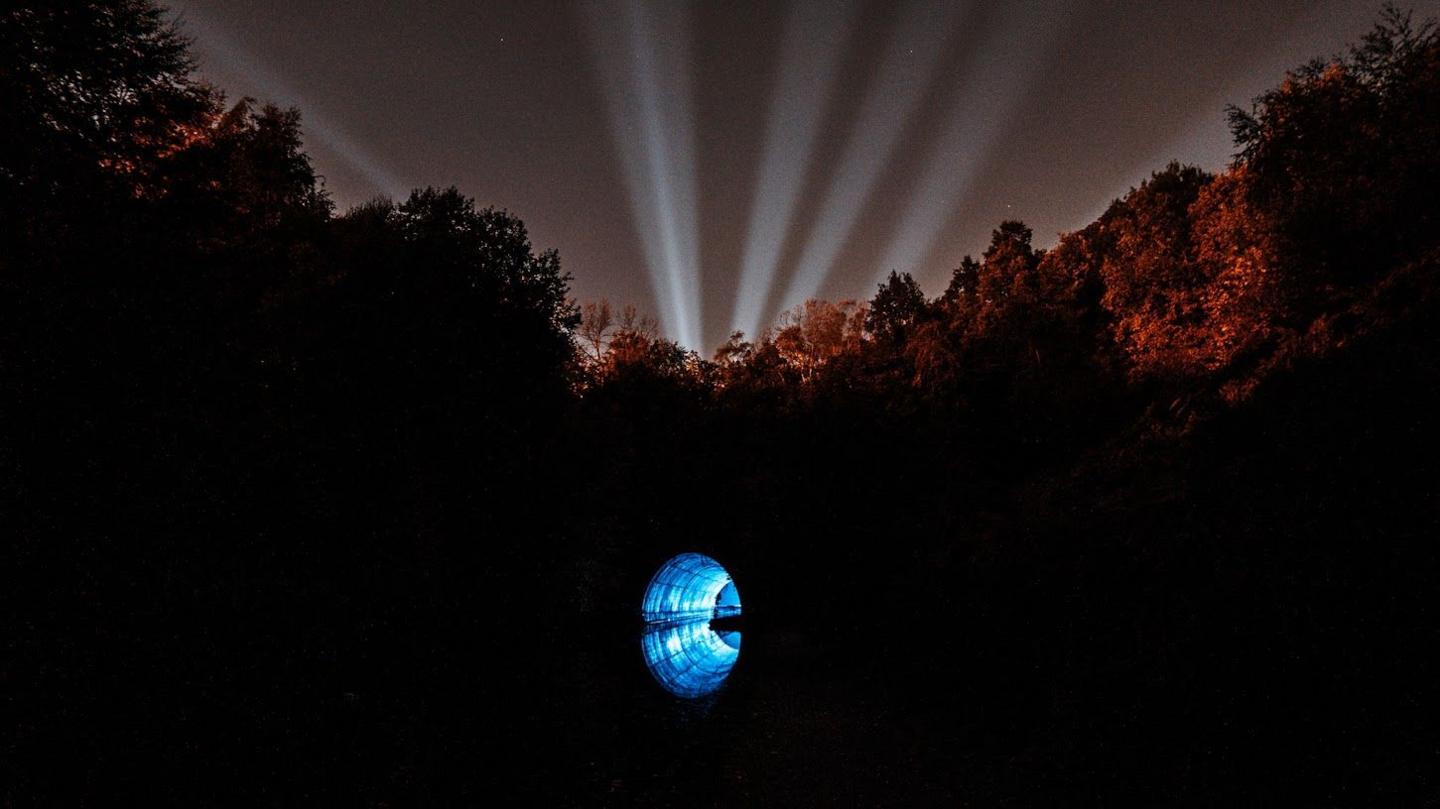 A canal bridge at night. The tunnel under the bridge is illuminated with blue light. The tops of the trees along the canal are also illuminated with red light.