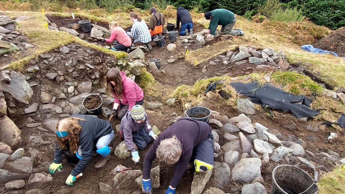 Archaeologists and volunteers carefully excavate a Neolithic site. They are using trowels and buckets.