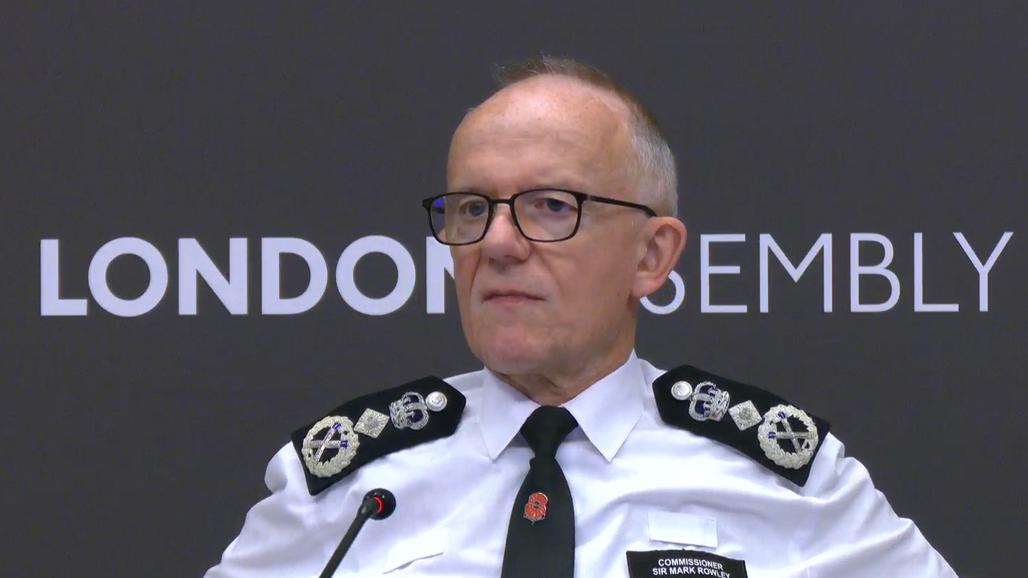 A medium-close up shot of Sir Mark in his police uniform, with black glasses on. He is straight-faced, looking to the left of the camera. He is stood in front of a London Assembly sign. 