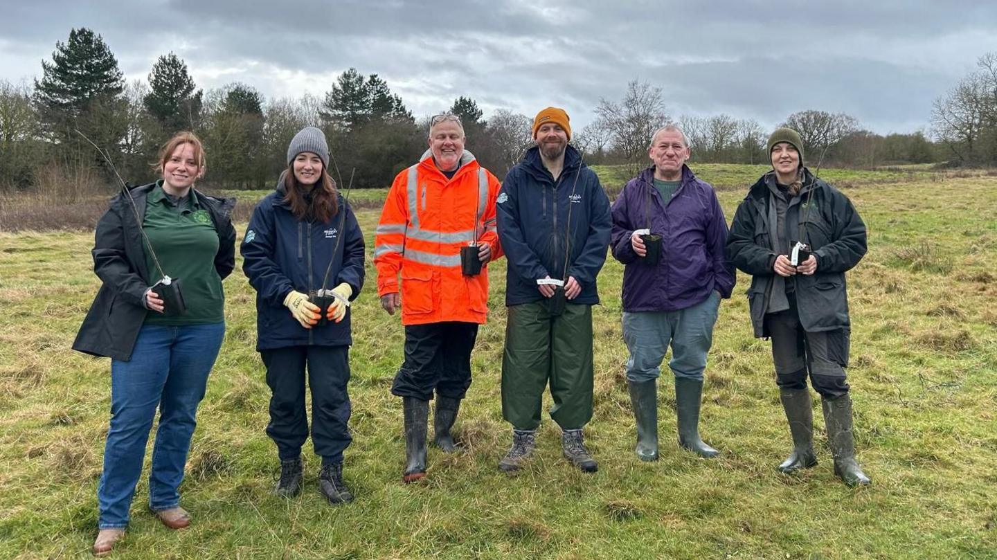 A group of volunteers gathered on a large area of grassland, holding saplings ready for planting, with a line of trees behind. They are all facing the camera and smiling.