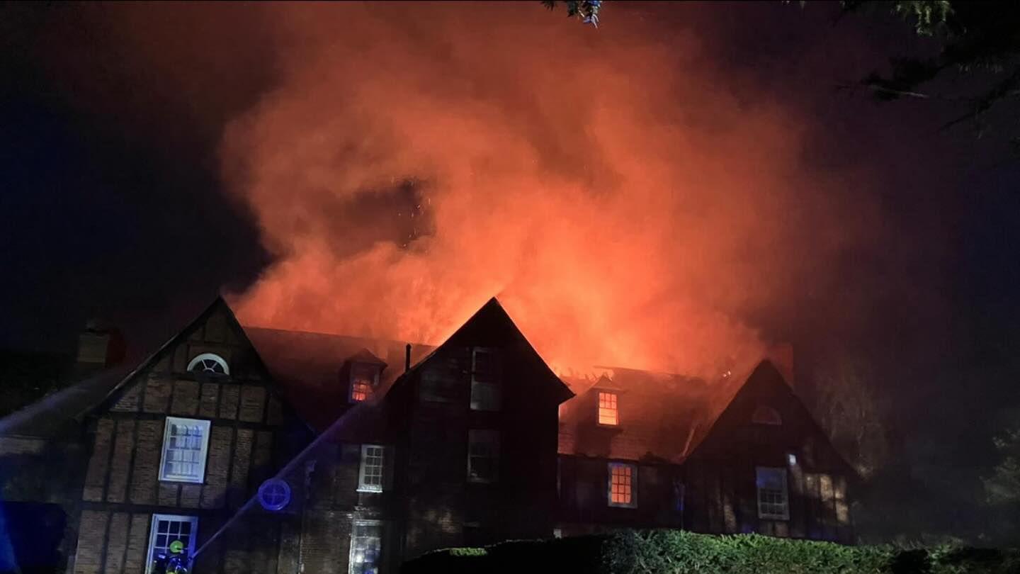 A night-time image of a large three-storey house. The roof is on fire and the smoke coming from he top the house is glowing red due to the blaze. In the centre of the fire the roof of the house has partially collapsed. 