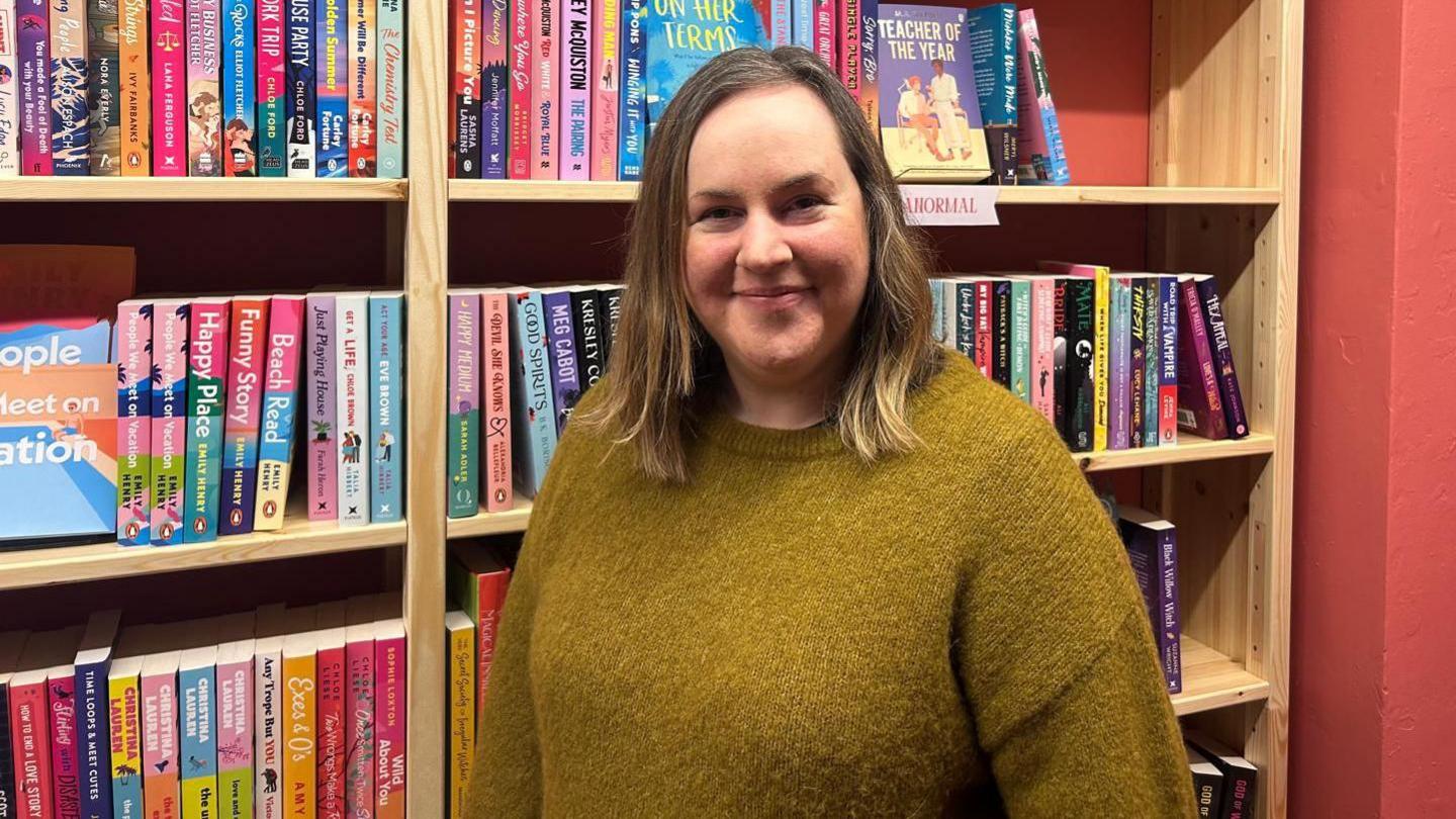 Llun o Angharad yn edrych ar y camera o flaen silff lyfrau 