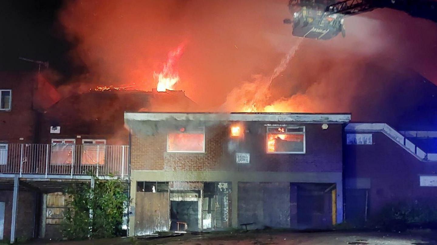 Red and orange flames can be seen behind and through the windows of a two-storey building. Just behind it is another two-storey building. A cherry picker device with firefighters on it can be seen above the building and a jet of water is going towards the flames.