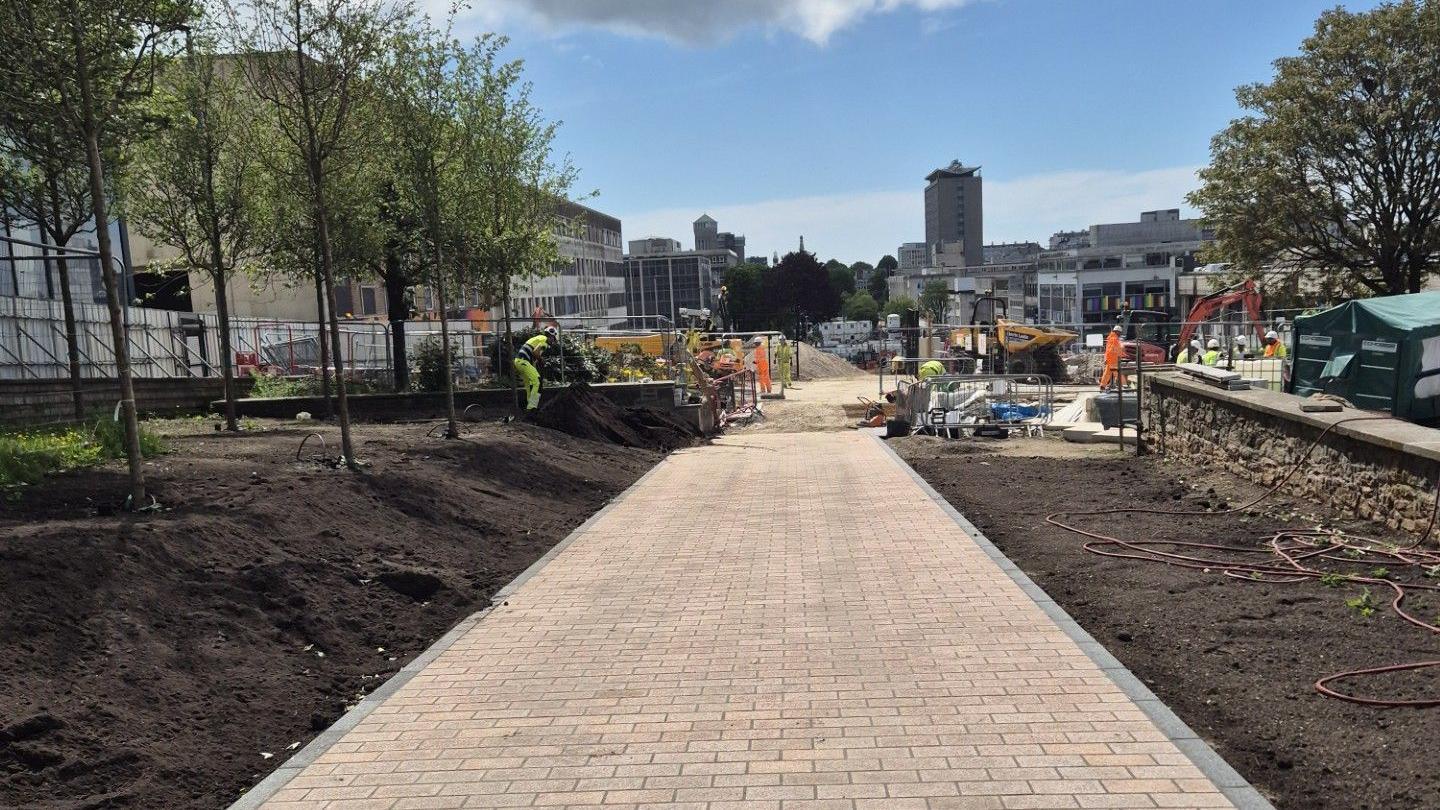Granite paving along Armada Way. Soil lines either side of the pathway. A worker, dressed in a hi-vis tabard, is stood on the soil. Trees have been planted. Machinery can be seen on the site. Plymouth's Civic Centre is in the distance.