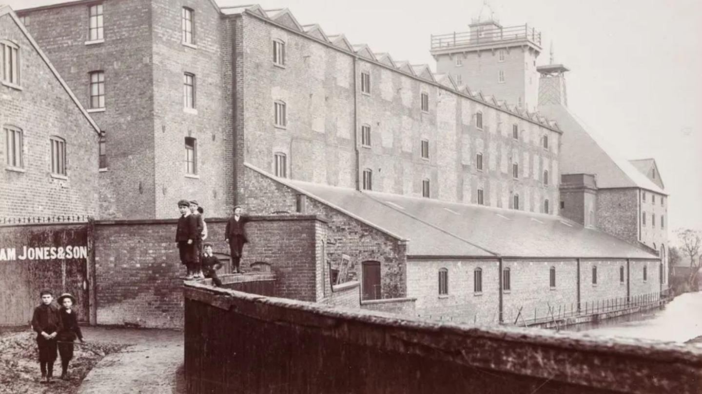 A black and white photo of a large, block-shaped, brick building with two towers on its roof and a number of children in dark Victorian-style clothing standing near the front