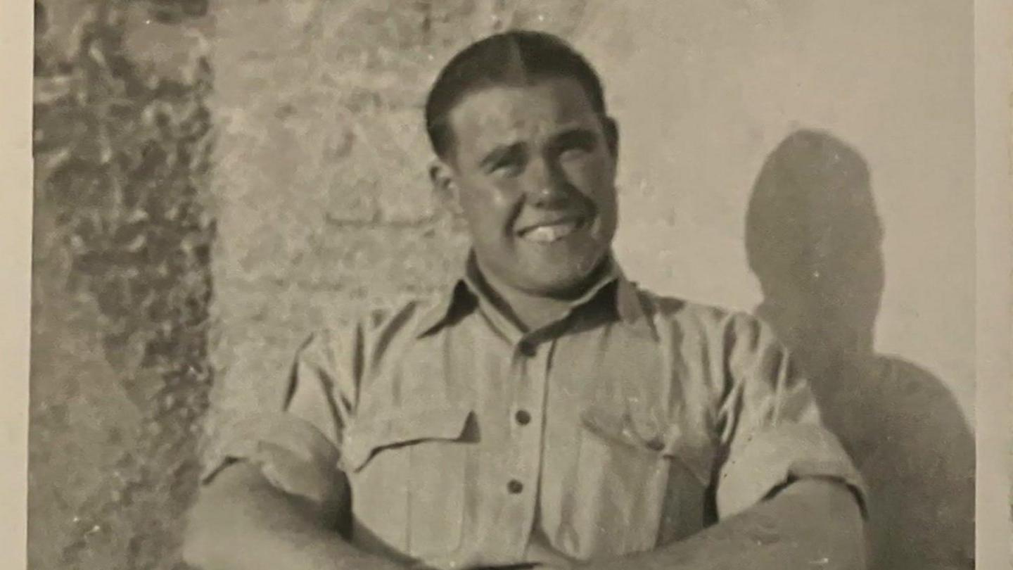 A black and white image showing Fred in a light shirt, sitting down and smiling as he looks at the camera.