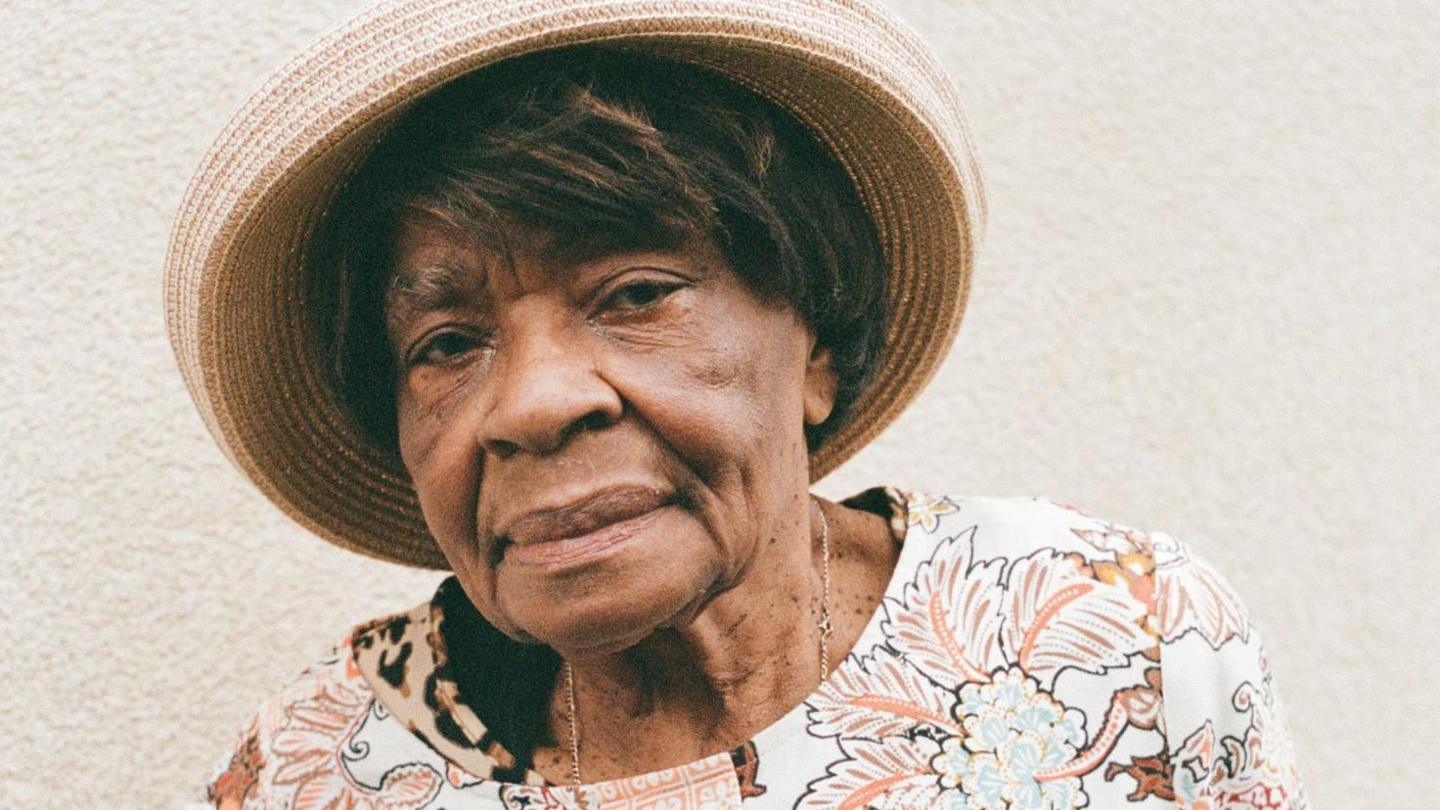 An elderly woman looking at the camera, wearing a straw hat and flowery top. 