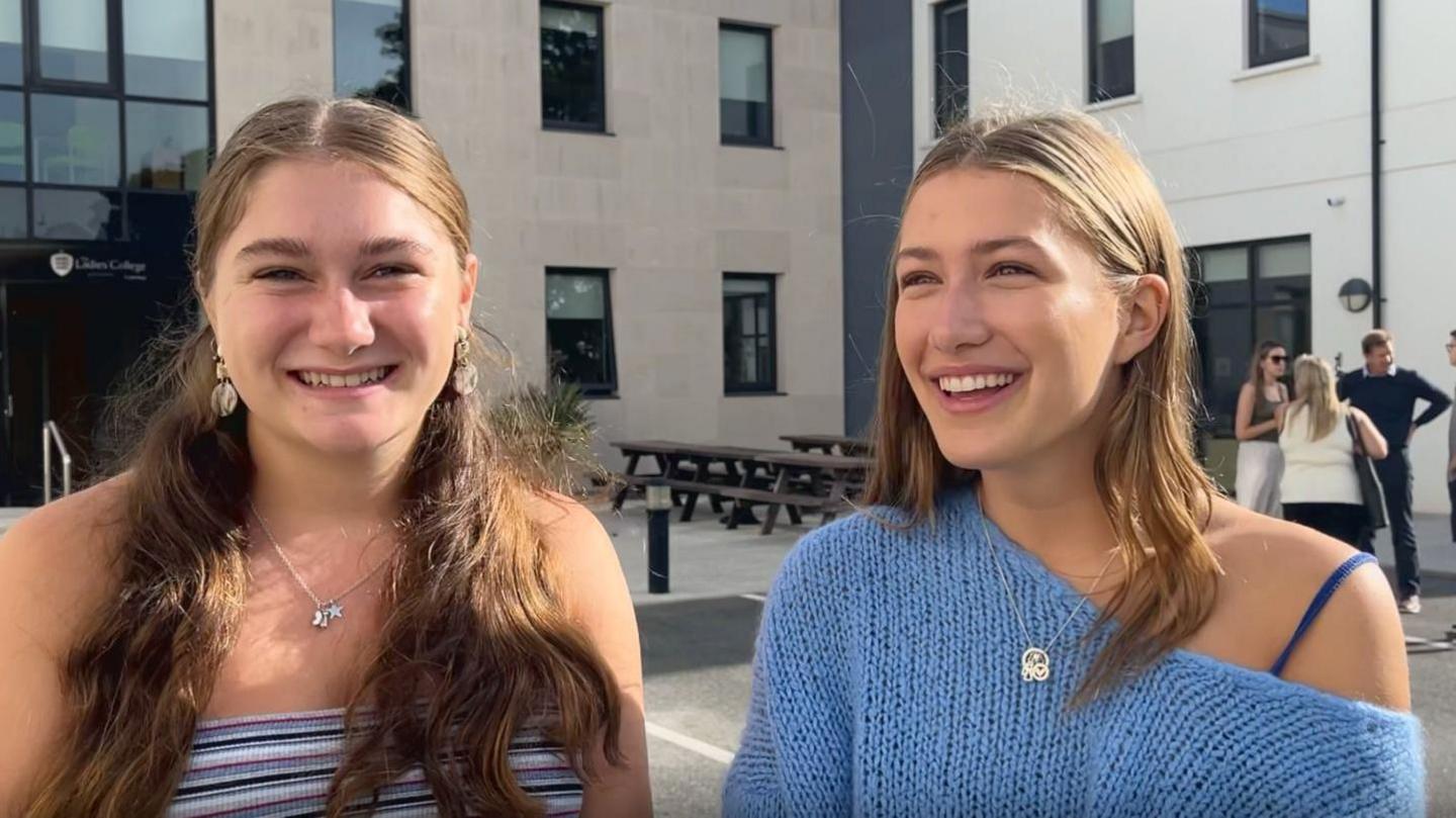 Poppy and Freya - Poppy on the left has long brown hair half up half down hairstyle, seashell dangly earrings smiling with teeth, wearing a  purple white and blue stripy strapless top, Freya with light brown hair smiling, wearing a light blue knitted long sleeve top off on one shoulder with a navy spaghetti stap, background of a grey building with rectangle windows, people speaking in a group to the right