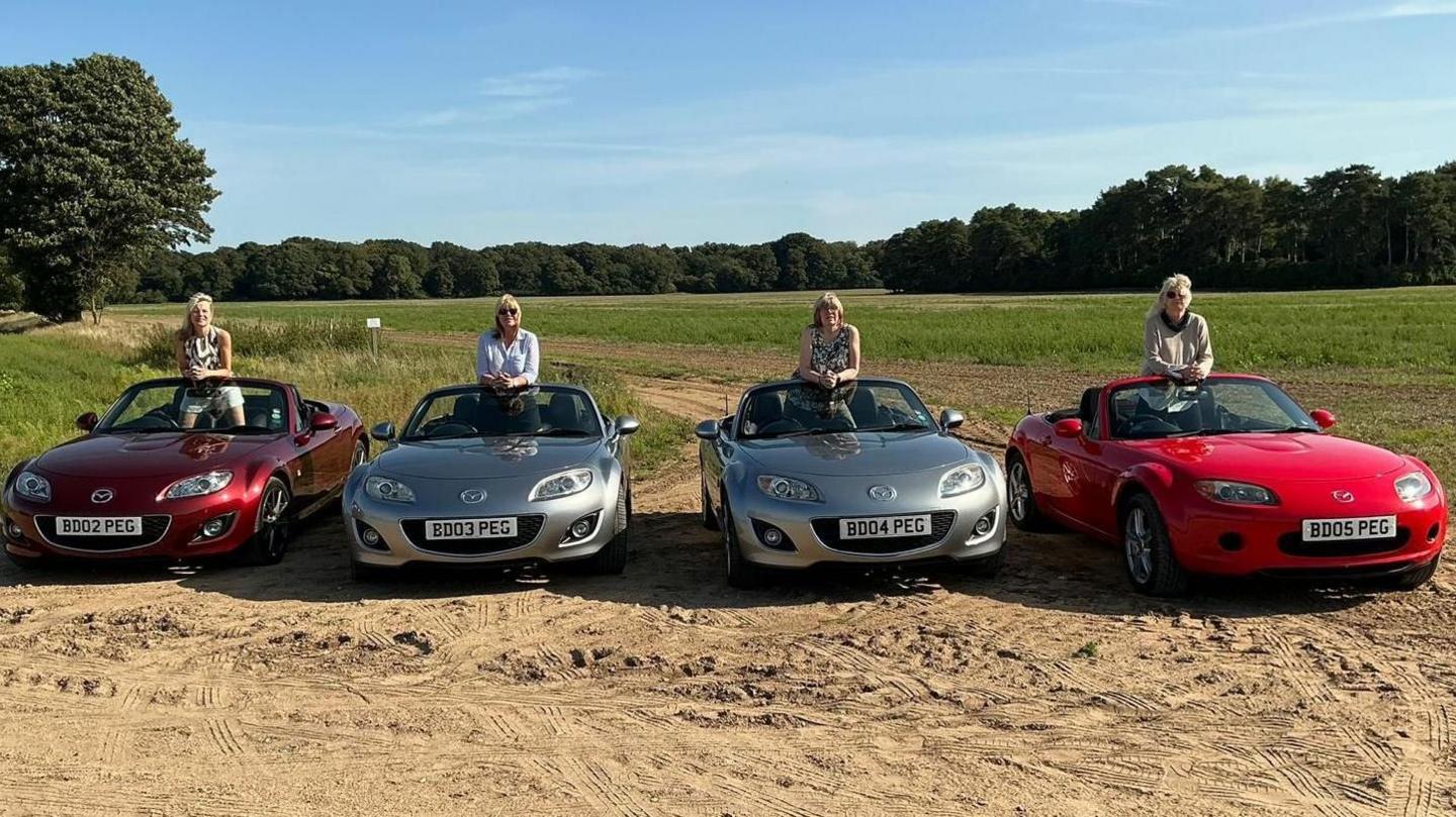 Four Mazda MX5 are lined up next to each other on a green field. The two on the ends are red and the two in the middle are silver. Each car has a person stood in the middle of it looking towards the camera.