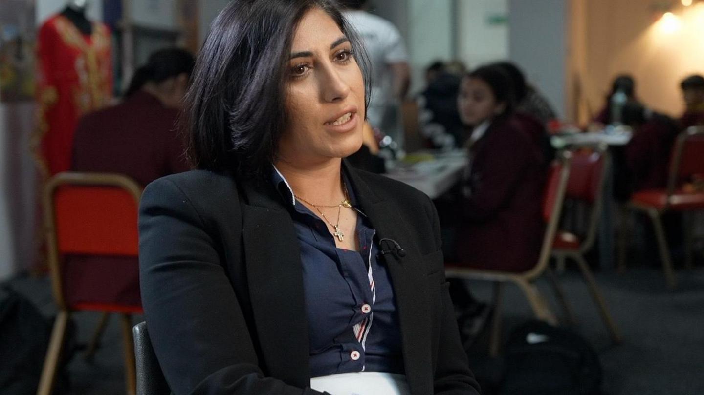 Irma Karchnakova is sitting on a chair. In the background, school pupils in uniforms sit around tables. 