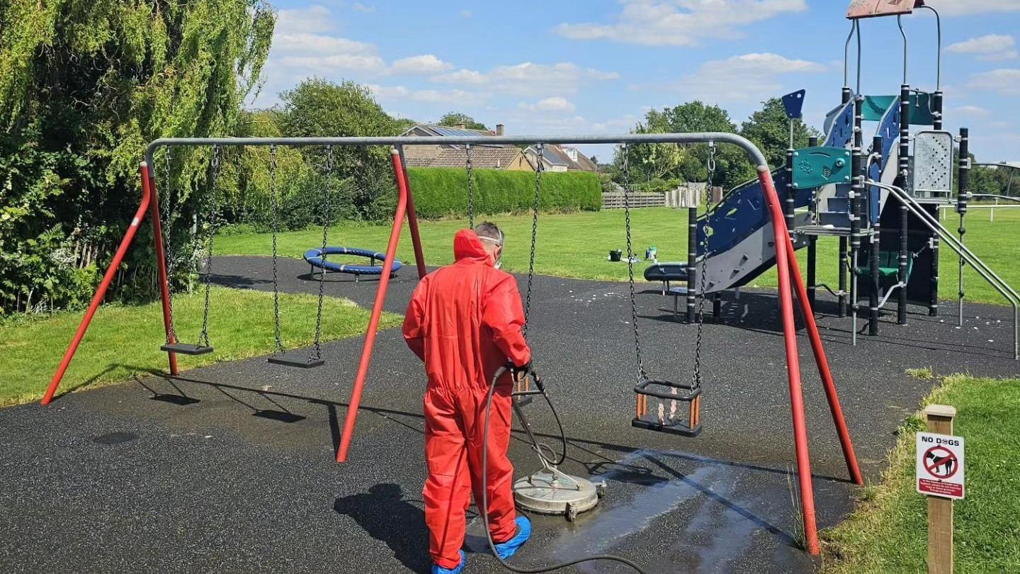 Man in red overalls sanitising the tarmac underneath a set of children's outdoor swings 