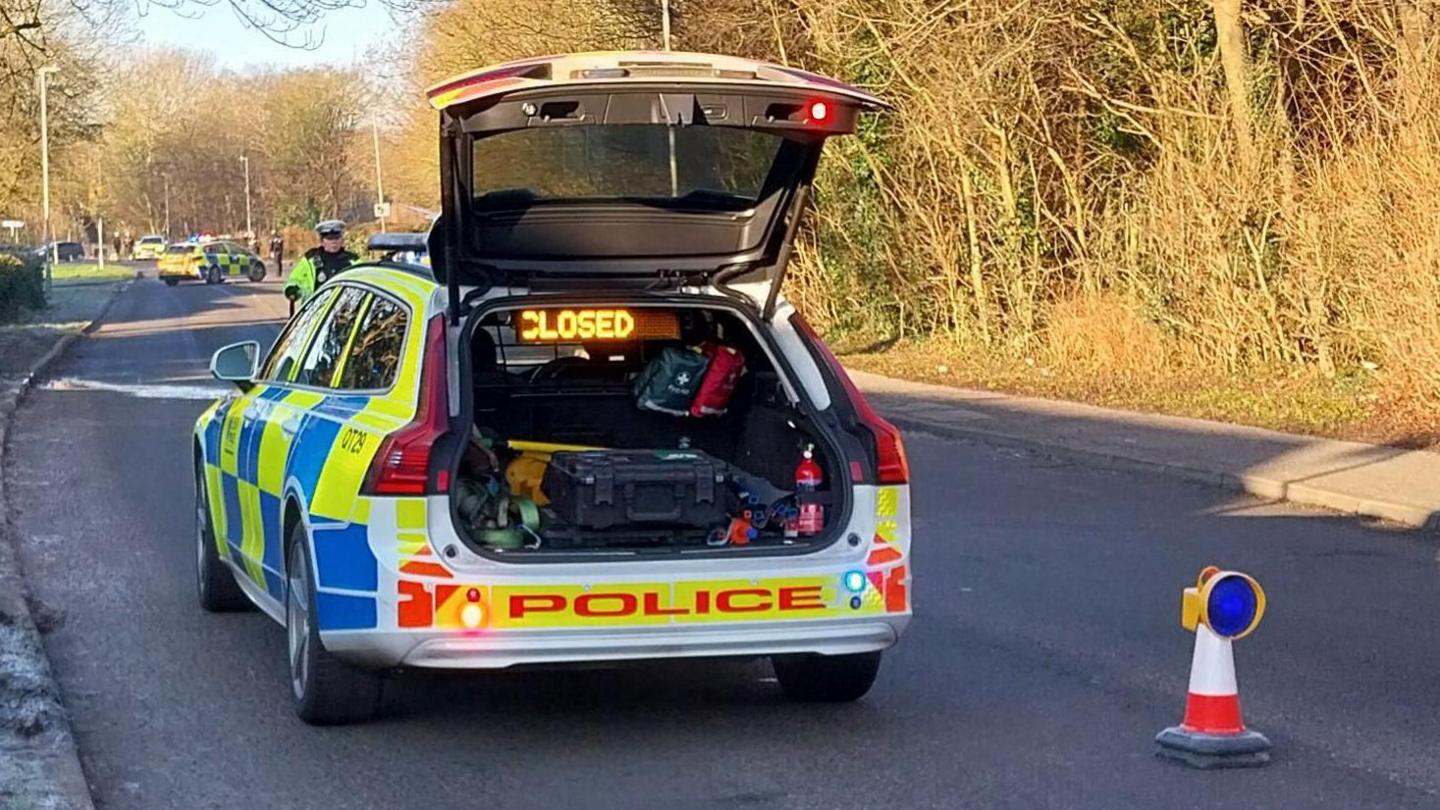 A police vehicle and cones blocking off a road. There is frost on the grass on a verge on on the left, and trees bathed in sunshine on the right.