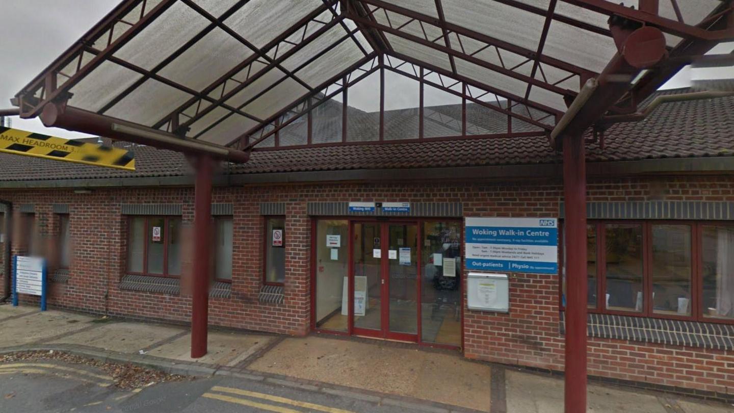 The outside of the Woking walk in centre which has a blue and white NHS sign on a red brick wall. There is a shelter with a glass roof over the door and all the metal work is a dark red.