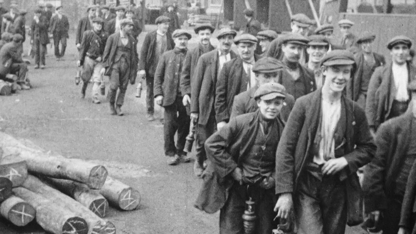 Men and boys dressed in flat caps and blazers and pants walking to work on a street. There are a pile of logs to the left and a long queue of boys and men to the right.