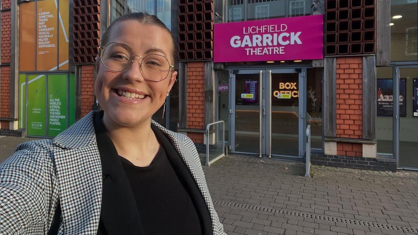 Rebecca is wearing a black top and a black and white blazer. She is standing in front of a brick building with a sign that says 'Lichfield Garrick Theatre'