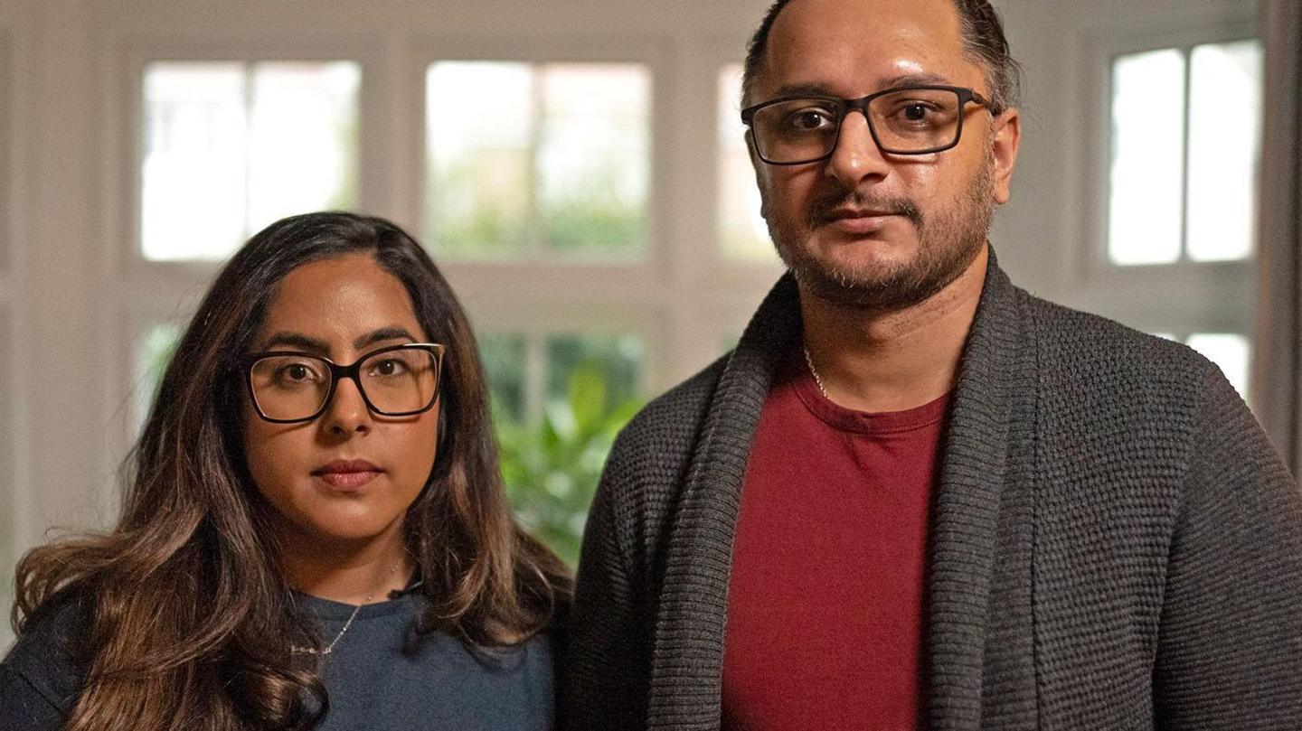 Amarjit and Mandip pictured standing next to each other. Amarjit has long brunette hair and black-rimmed glasses. She is wearing a blue jumper and a silver necklace. Mandeep has dark hair which is tied back, black-rimmed glasses and a short beard. He is wearing a red t-shirt and grey woollen cardigan. They are pictured in front of a white-framed window with green plants outside.
