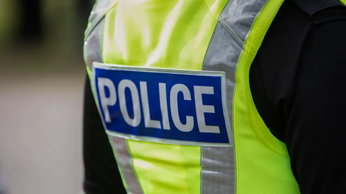 The back of a police officer with a black long-sleeved top on, and a yellow hi-vis jacket with silver reflective strips. It says "police" on a blue background in silver writing.