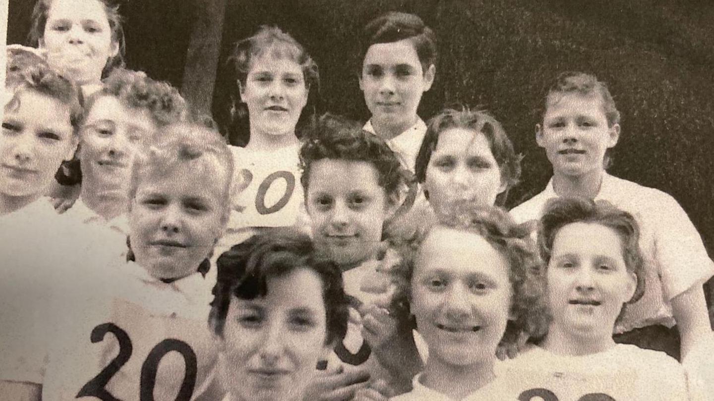 A black and white photos of 12 schoolgirls looking towards the camera. They are wearing white sports shirts, some with the figure 20 pinned to the front. 