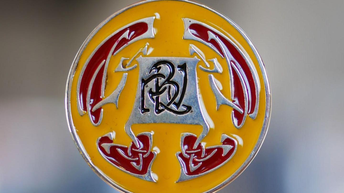 A closeup of the RBV medal, which is silver with a yellow and red Celtic design around the letters RVB in black in the middle.