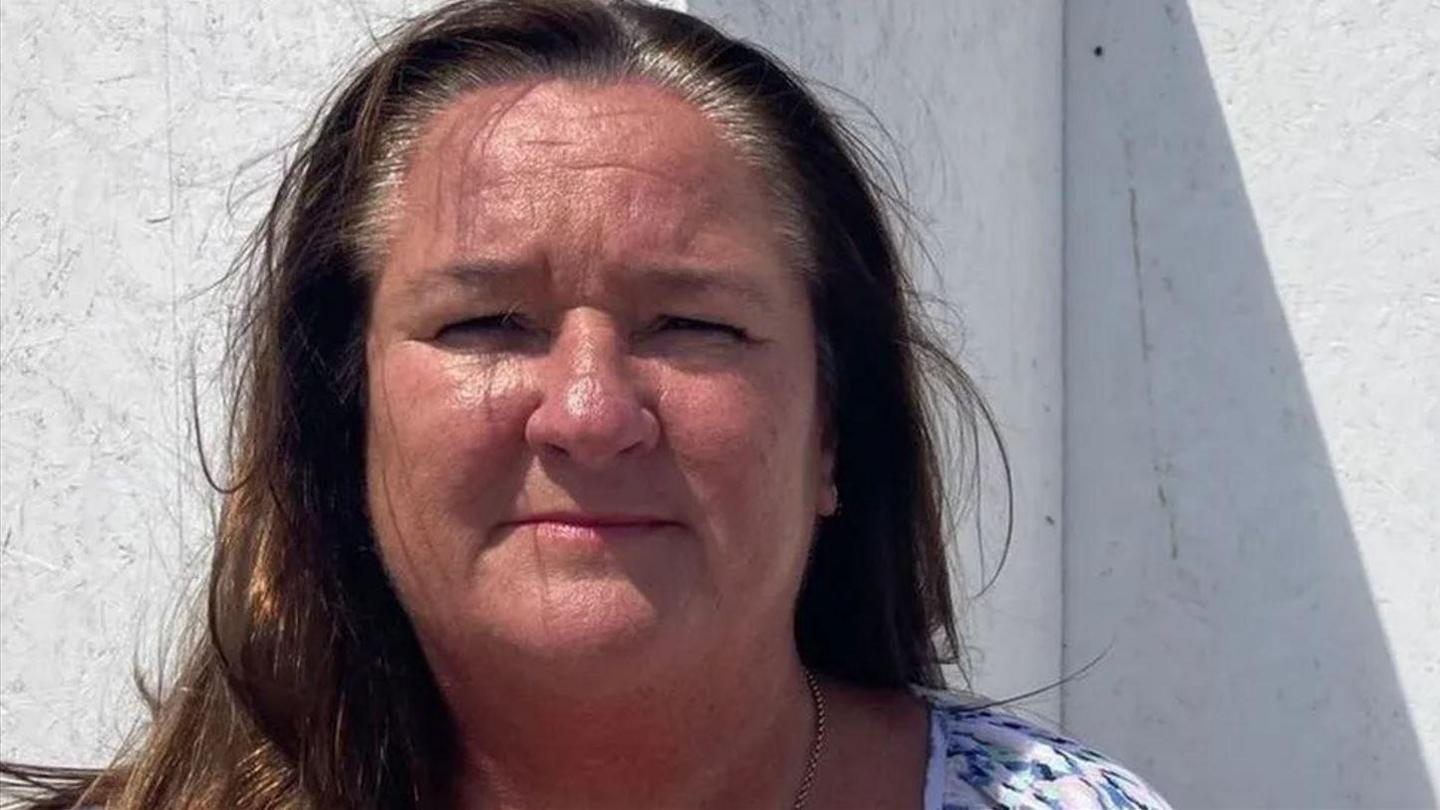 Marion Atkinson squints into the camera in the sunlight. She has dark brown shoulder-length hair and is wearing a necklace.