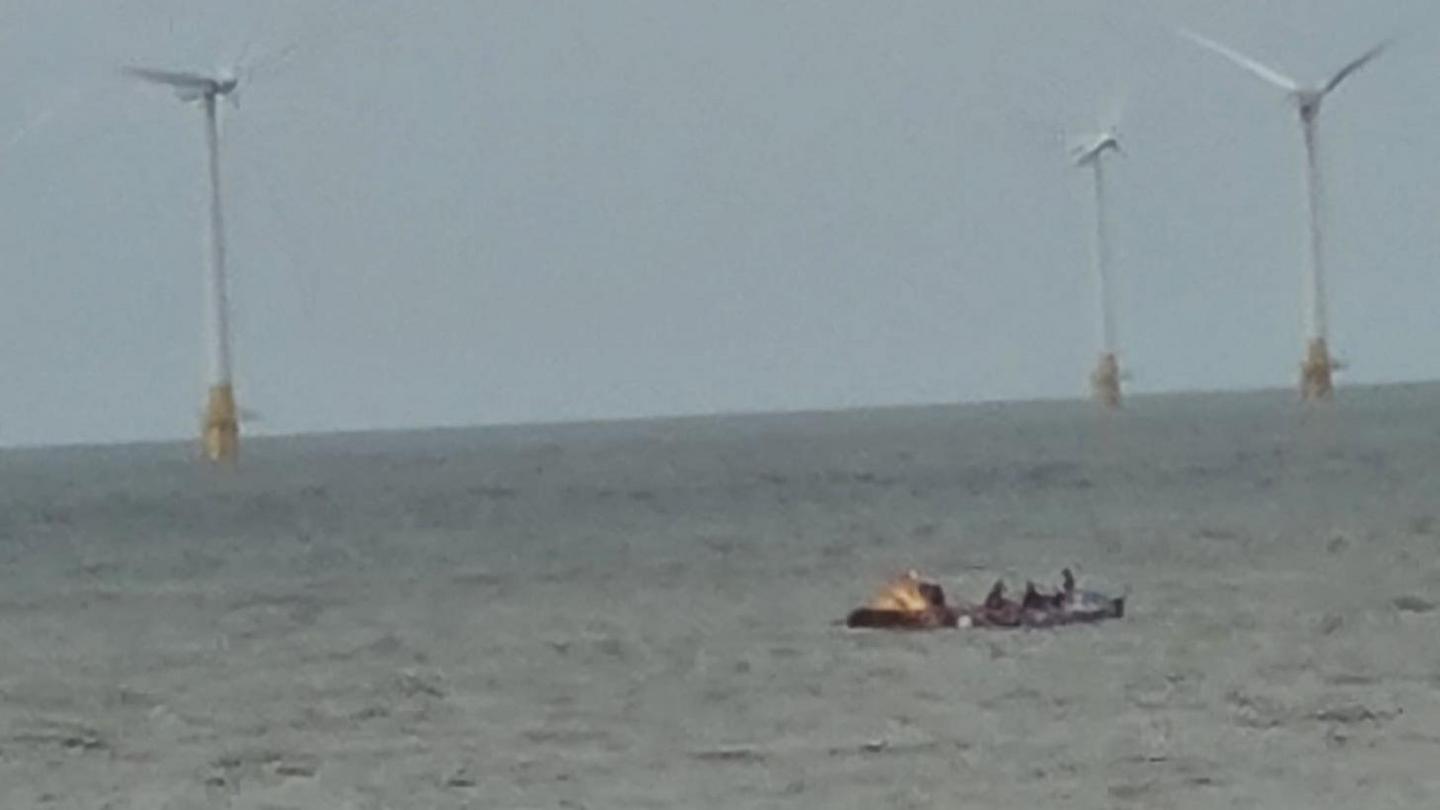 A picture taken by Rupert Lowe of a boat and rowing crew out at sea. In the distance there are three wind turbines. 