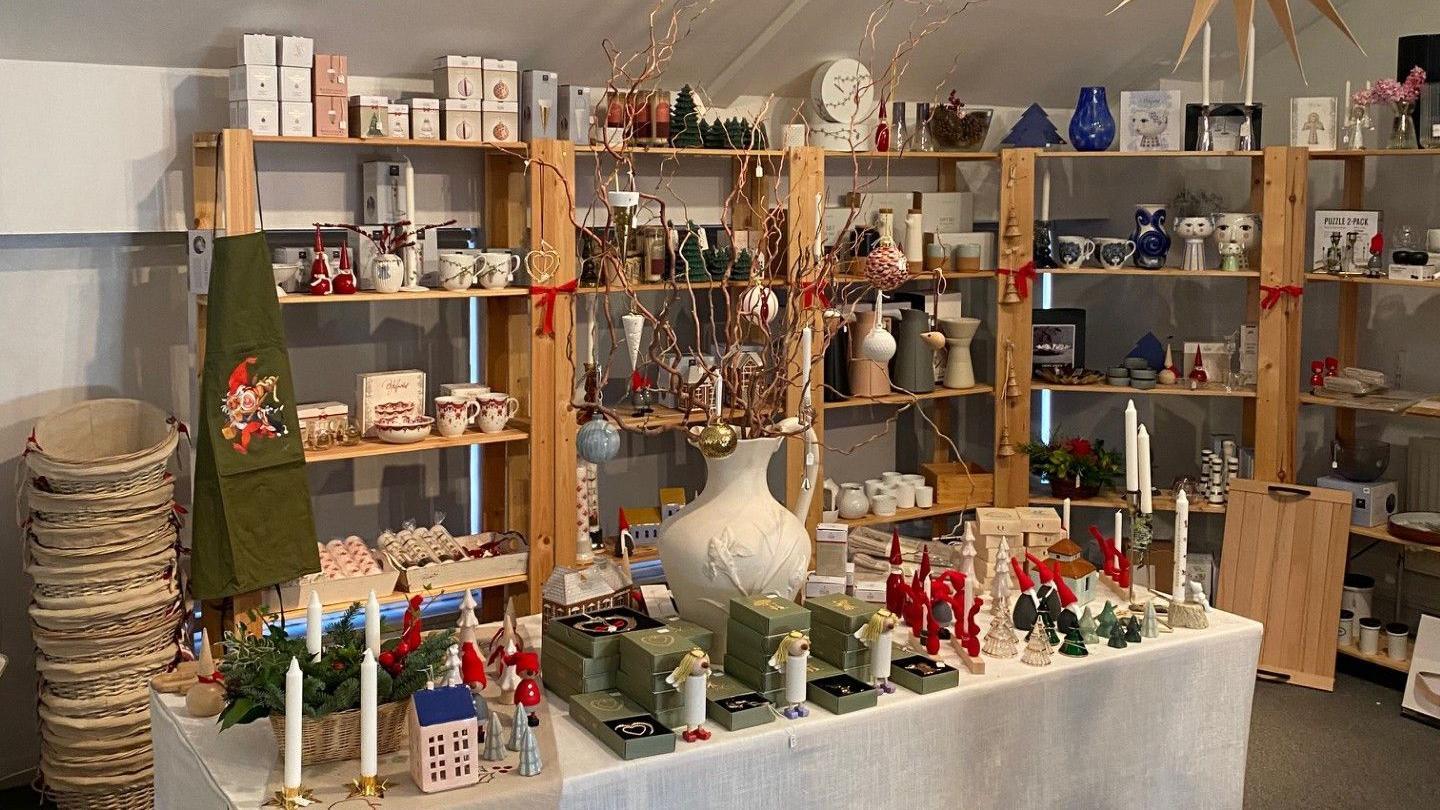 A table with a white tablecloth on and a collection of candles. There are shelves behind it that are full of candles. 