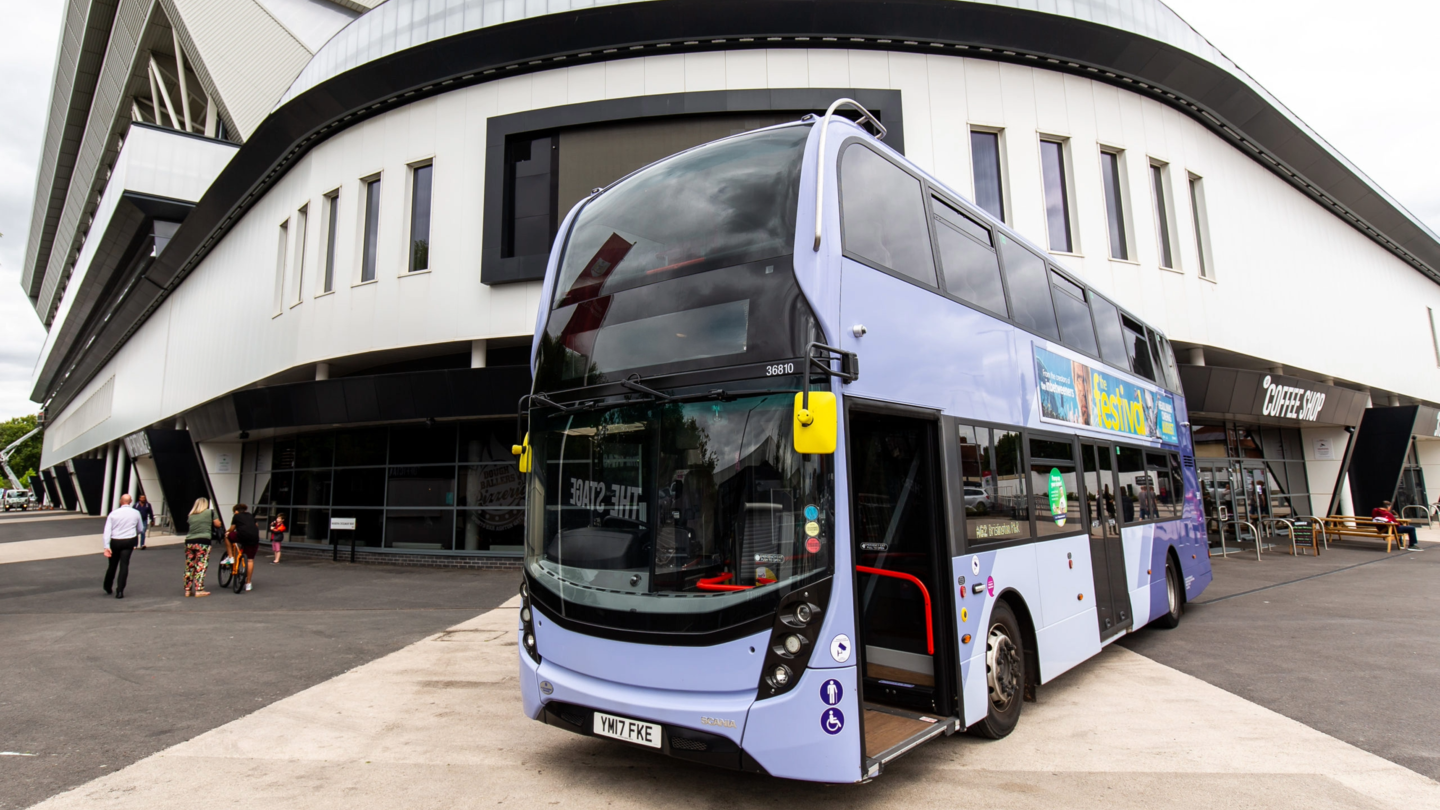 A purple bus is parked outside Ashton Gate Stadium. There are people standing around in the distance and a coffee shop sign to the right.