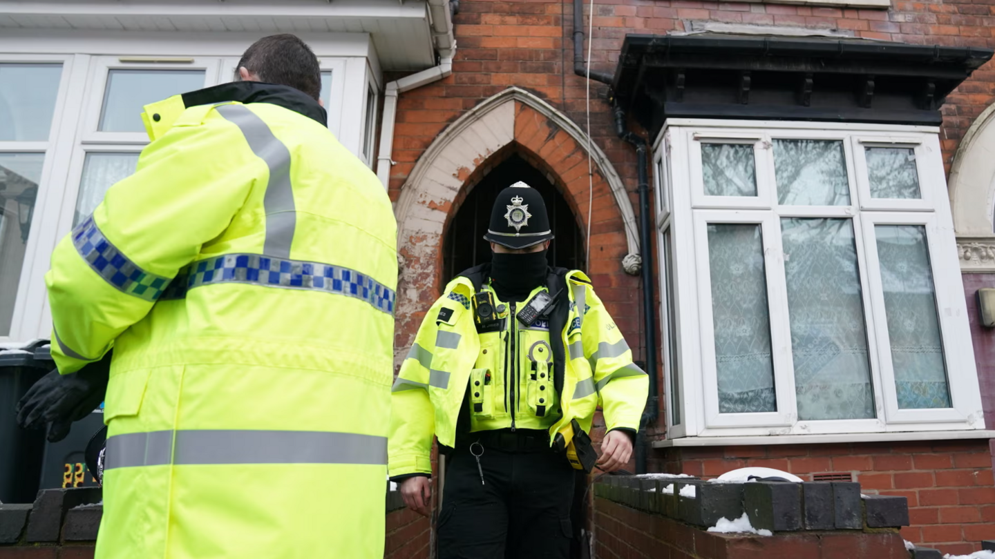 Two police officers leaving a red brick house with white windows. Both officers are wearing fluorescent jackets, with one in a police had and a facemask