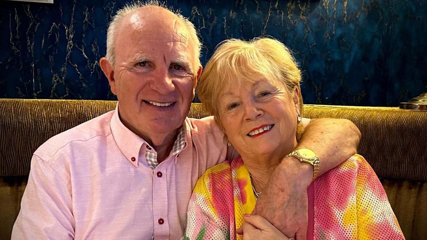 Gabriel and Josephine Dolan sit together smiling. Gabriel is on the left and has his arm around Josephine's shoulder. He is wearing a pink shirt and gold watch. Josephine is wearing a multi coloured top, silver earrings and has blond hair.