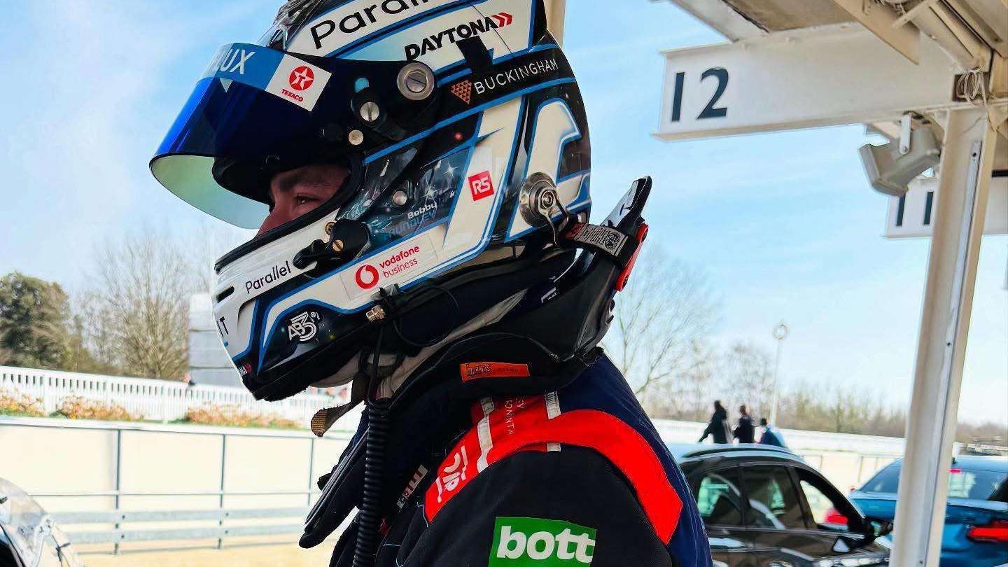 A race car driver in full gear stands beside a parked car in the pit lane, wearing a helmet with "Parallel" and “Daytona” branding, under a shelter marked with number 12.