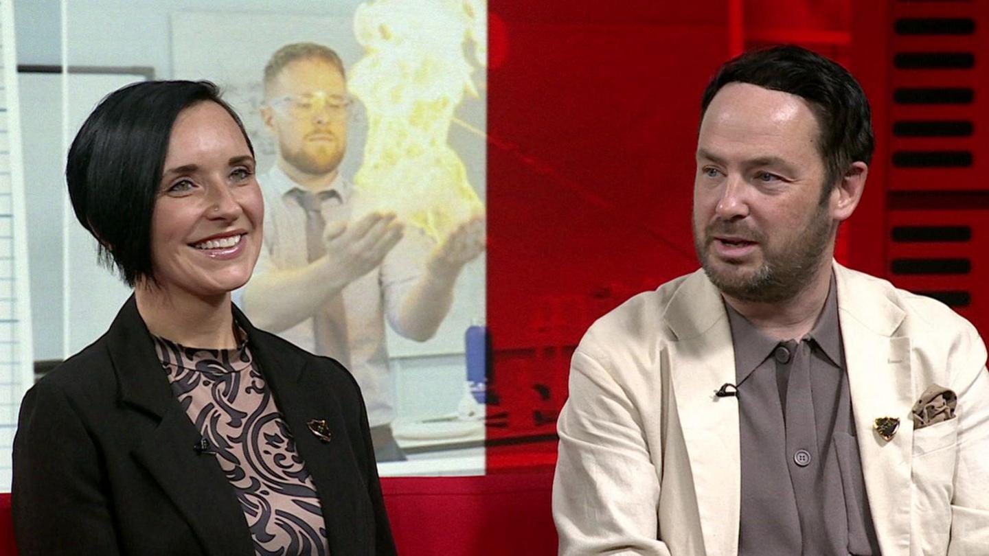 Two individuals sit on a red couch, facing slightly to their left. The person on the left wears a patterned dress with a dark blazer, and the person on the right wears a light-colored jacket over a shirt. Behind them is a backdrop featuring an image of someone conducting a fire-based experiment, along with graph paper and office-themed visuals.