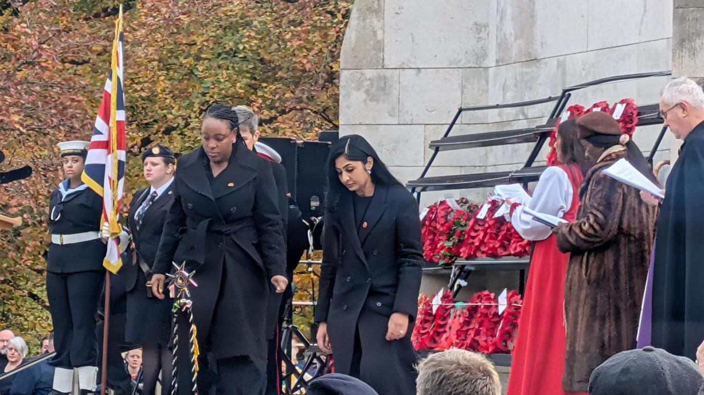 Two women with dark hair and dark coats standing with their heads bowed with women in military uniform to their right, holding a flag and an array of poppy-wreaths to their left