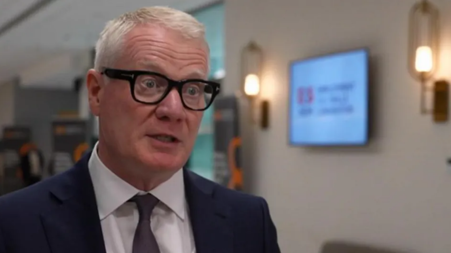 Richard Parker in a dark blue suit, with a purple tie and white shirt and glasses. The background is out of focus but a TV is visible mounted to the wall, between two light fittings