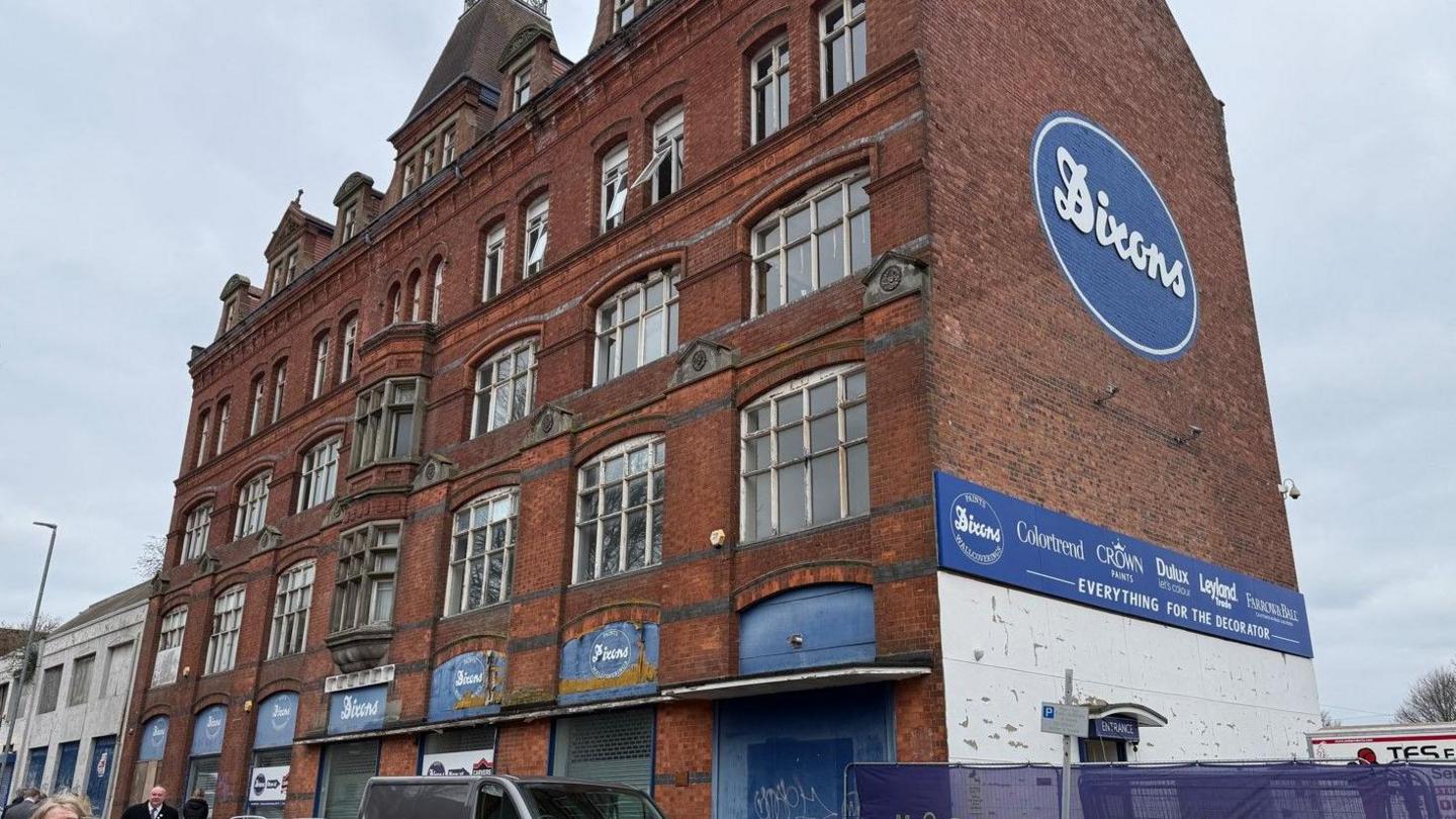 A five-storey building photographed at an angle. The Dixons logo, white words on a blue background, is in various places, with the largest version on the side of the building.