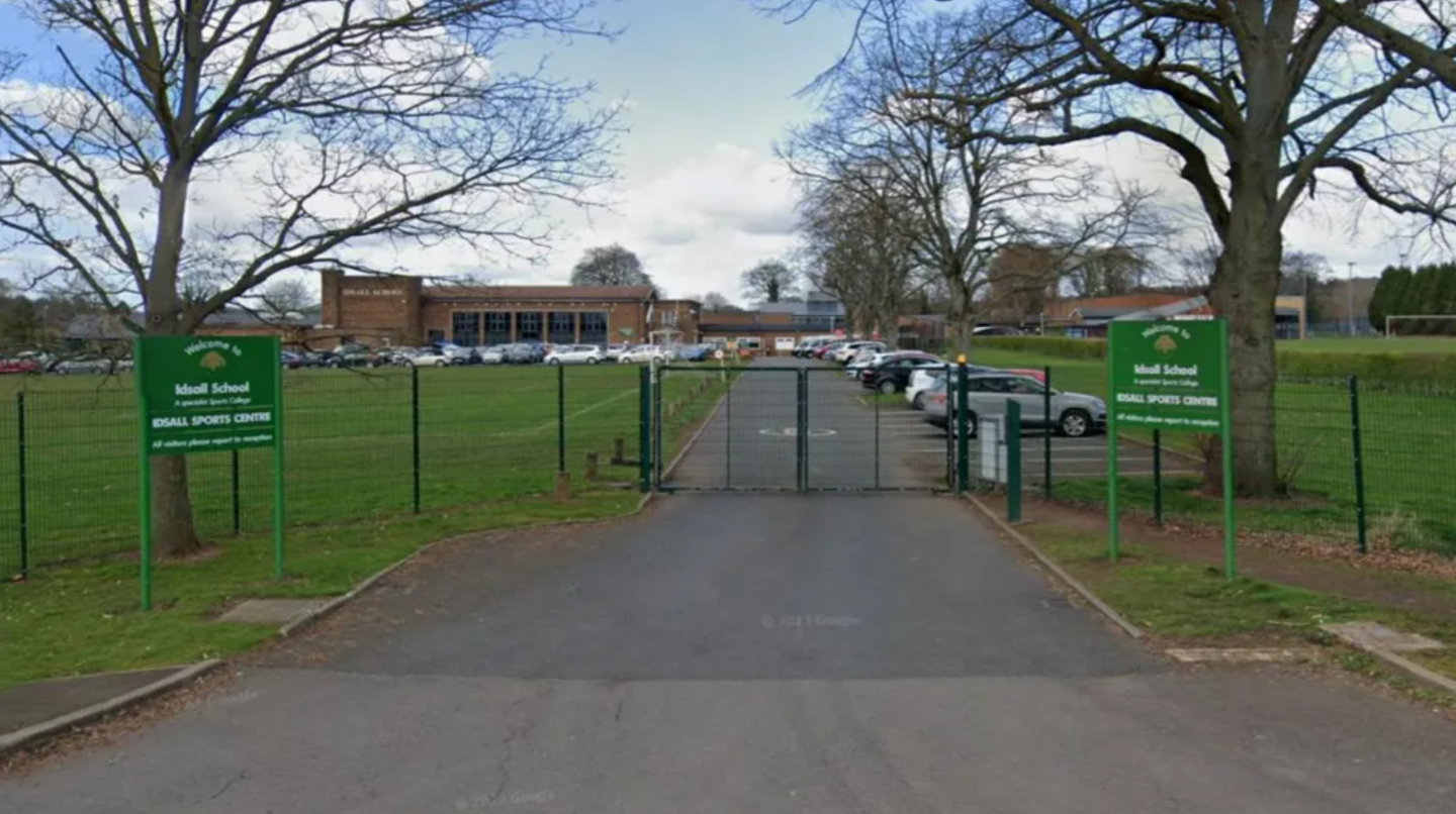 The entrance gateway to Idsall school with a metal gate in the middle of a drive and two green signs on either side