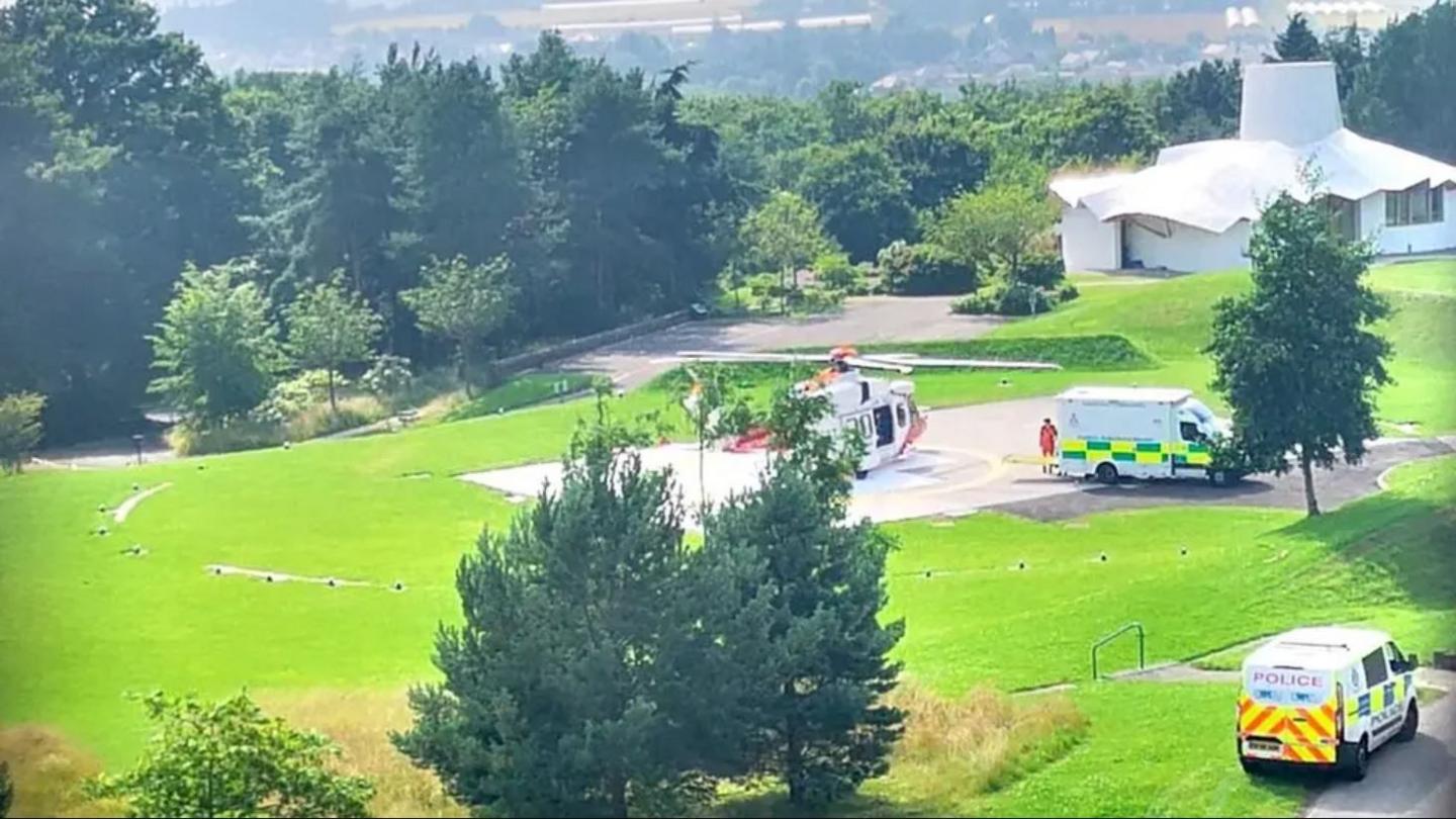 A helicopter pad with a helicopter near Stanley surrounded by trees. An ambulance and a police van are also in the picture