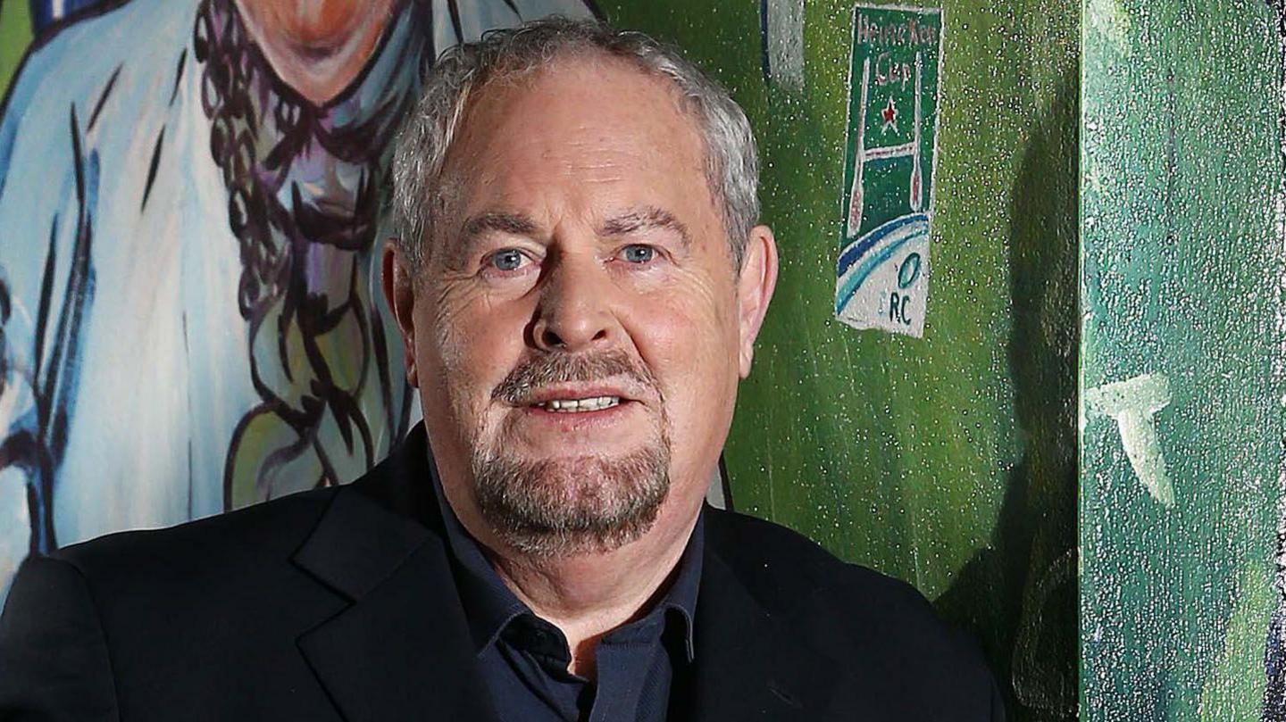 A man with short white hair looks at the camera. He is wearing a dark shirt and a black blazer. He is standing in front of a green wall.