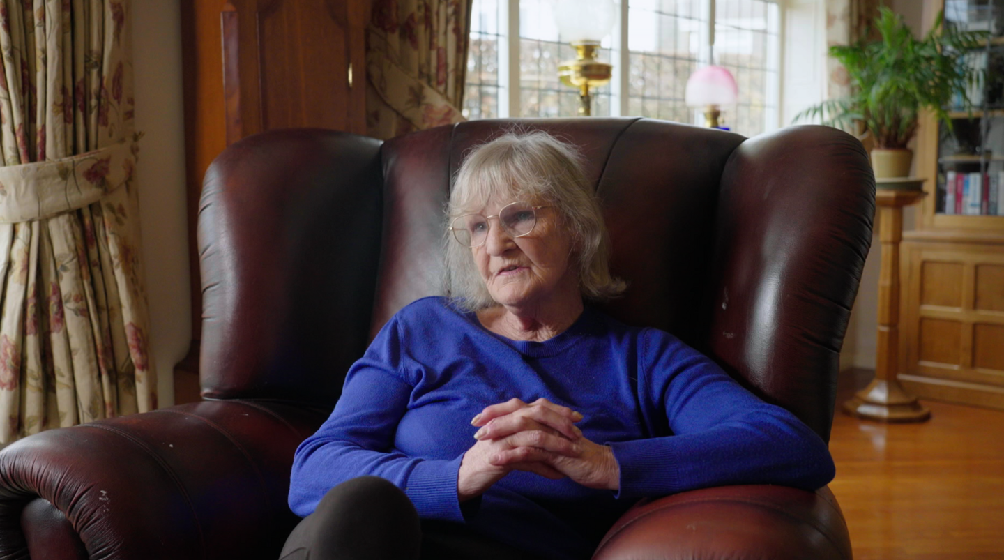Mary Lanigan, the former leader of Redcar and Cleveland Council, sits in a large, reddish-brown leather armchair. She wears a blue long sleeve top and glasses. She has shoulder length grey hair and sits with her legs crossed and her fingers linked together on her lap.