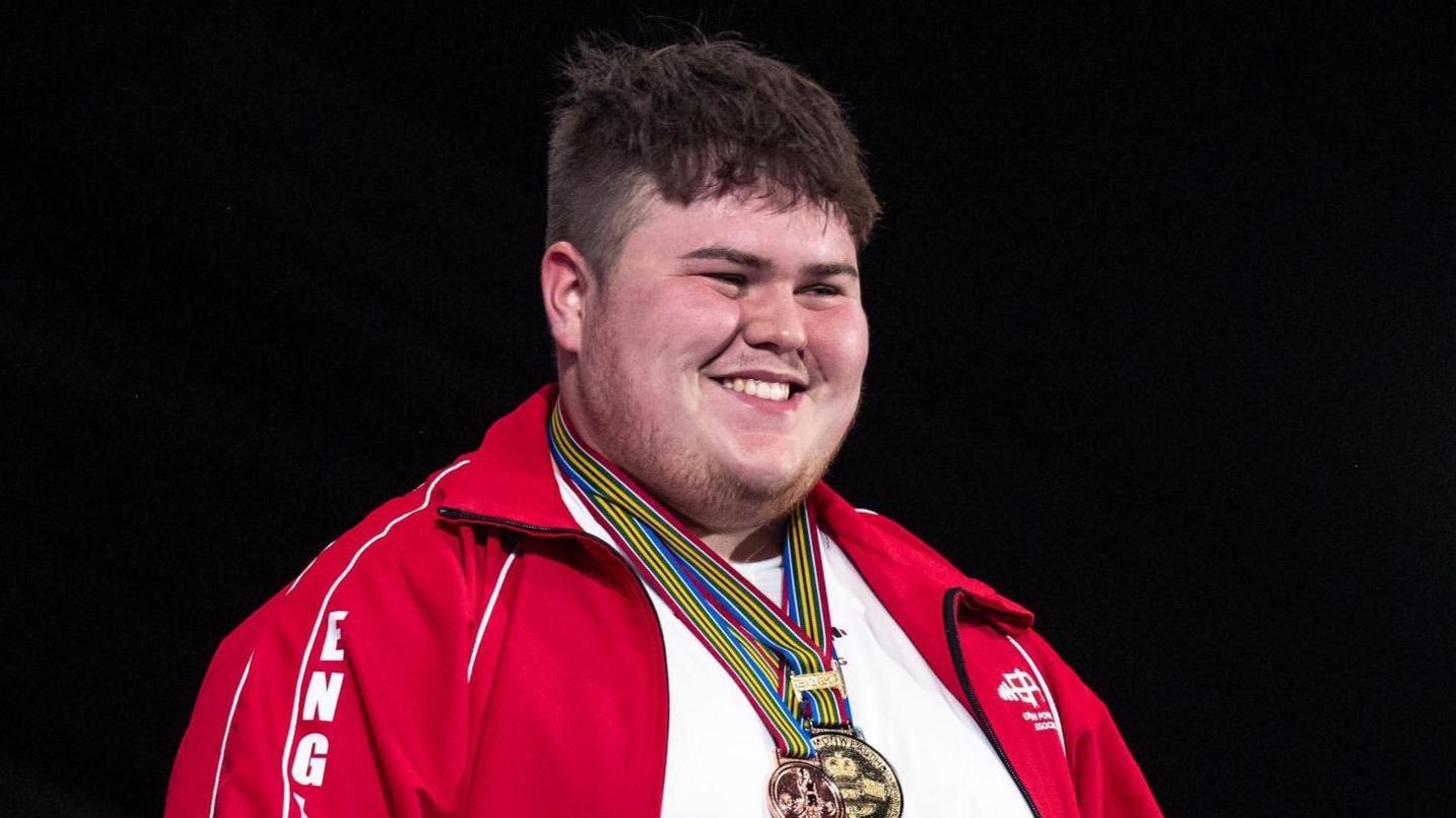 Cleverley smiles and looks away from the camera. He has two medals around his neck and he wears a red jacket with England written on one arm. Underneath he wears a white top. 