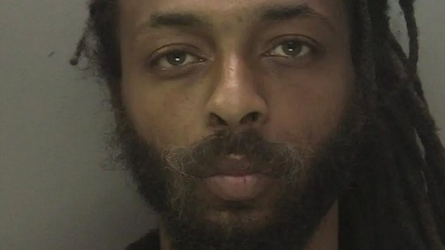 Ishmael Farquharson  with a blank expression on his face. He has various length locs and a full dark beard.