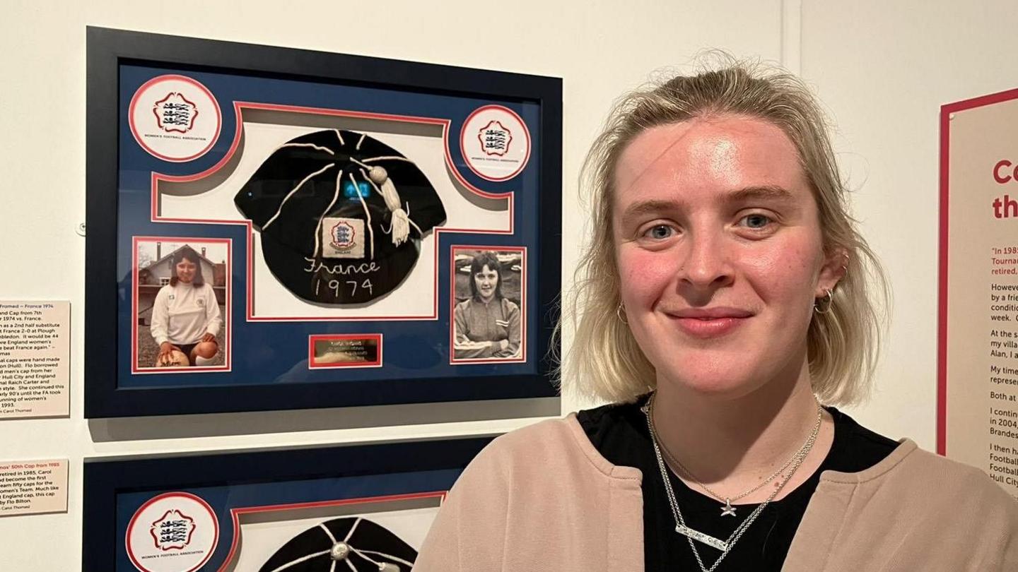 A young woman with blonde bobbed hair stands next to a display board featuring handmade black caps with tassels on. She is smiling. 