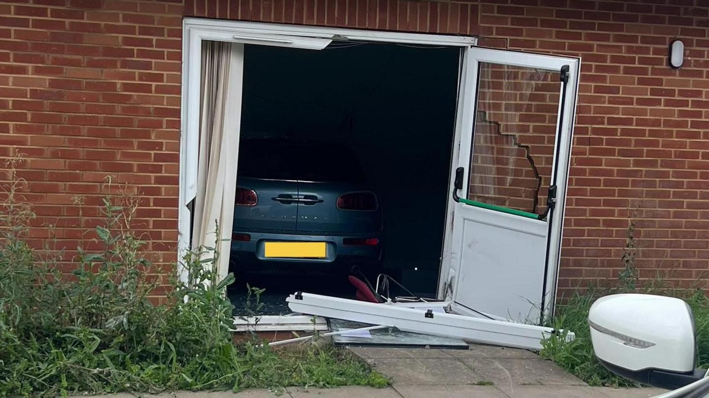 The rear view of a grey car which has crashed through French windows. It has crossed the threshold and is fully into the room. One door is open on the right and the other is in pieces on the ground in front of the brick built building.