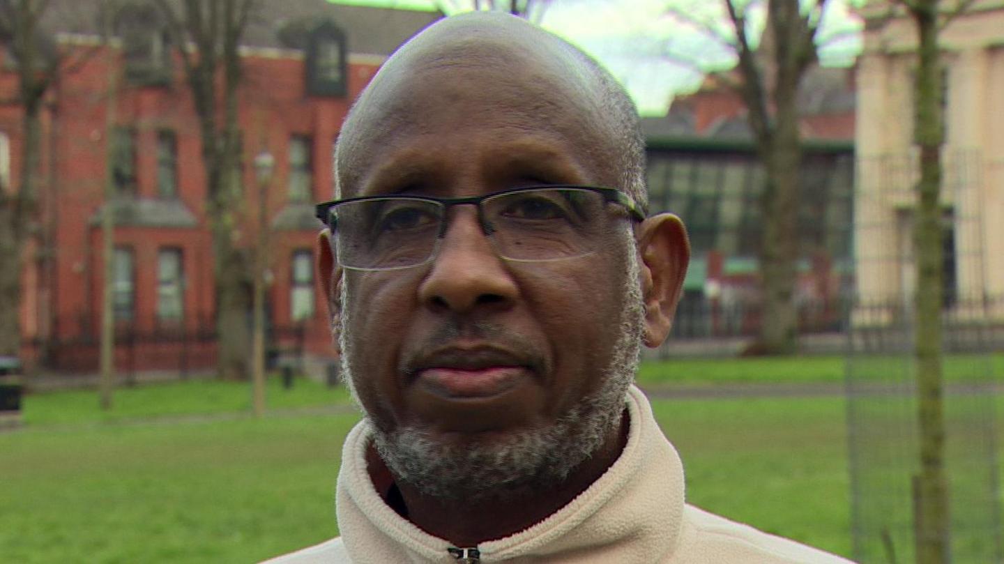 A man is standing in an open area with grass and trees. He's wearing a white hoodie and glasses.